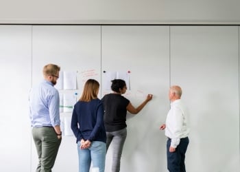 Pessoas reunidas em uma sala de reunião enquanto uma delas escreve ideias em um quadro branco durante uma sessão de planejamento colaborativo.